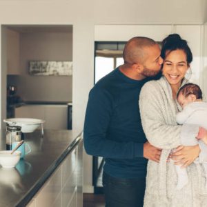 Man kissing his wife holding a newborn baby boy in kitchen. Lovely young family of three in morning in kitchen.