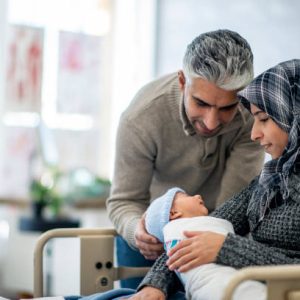 A mother and father are indoors in a hospital room. The mother is wearing a head scarf, and she is holding her newborn baby boy. The father is touching his son and watching him sleep.