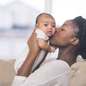 A beautiful young African American mother gently holds her infant daughter up in the air with both hands and kisses her cheek. The baby's eyes are wide open and she looks happy. They are sitting on a couch in their living room.