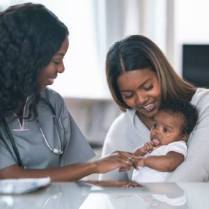 A beautiful Indian woman is holding her baby at a medical appointment. She is sitting next to the pediatrician at a table in a medical consultation room. The doctor is a black woman. The doctor and mother are both smiling while looking at the baby.